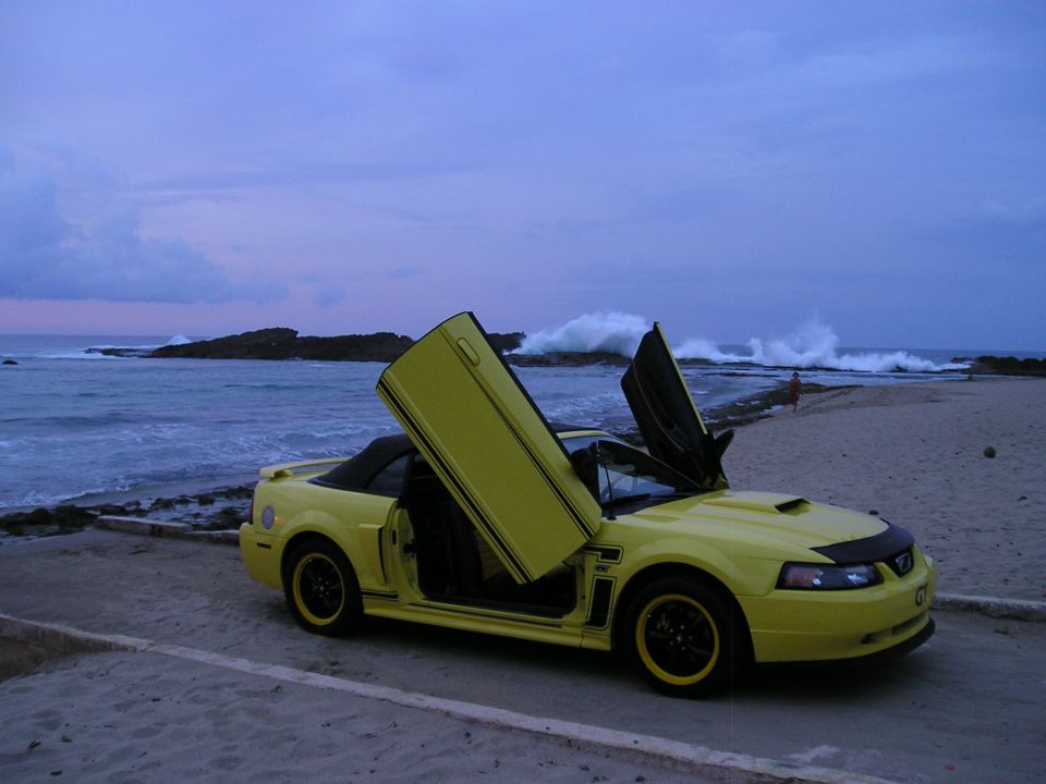Yellow convertible Mustang with doors open on a beach at dusk.