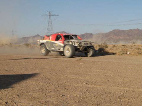 1996 Ford Bronco - A red off-road race truck navigates a dusty desert track, its front end partially obscured by dust.