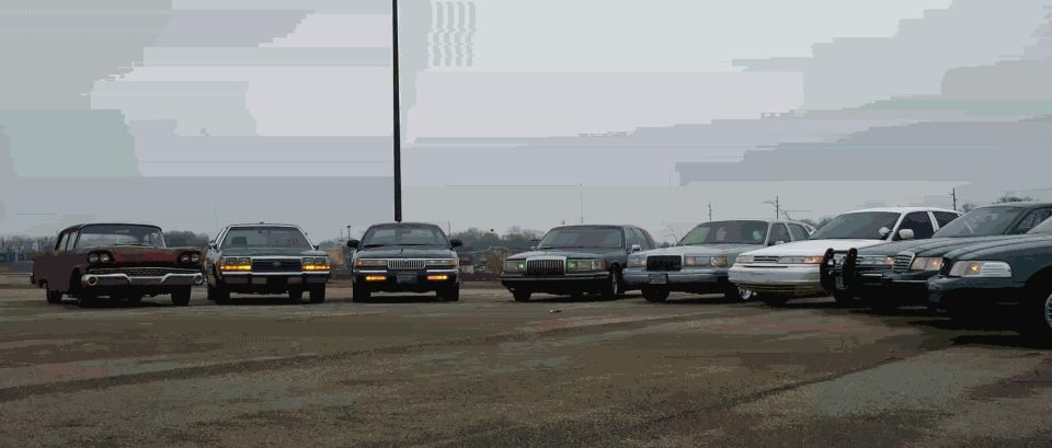 Curved lineup of full-size sedans facing camera on a gravel lot, rusty 1950s Ford at left