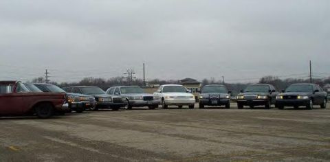 1988 Ford Crown Victoria - Several sedans and a pickup truck are parked in a line on a gravel lot.