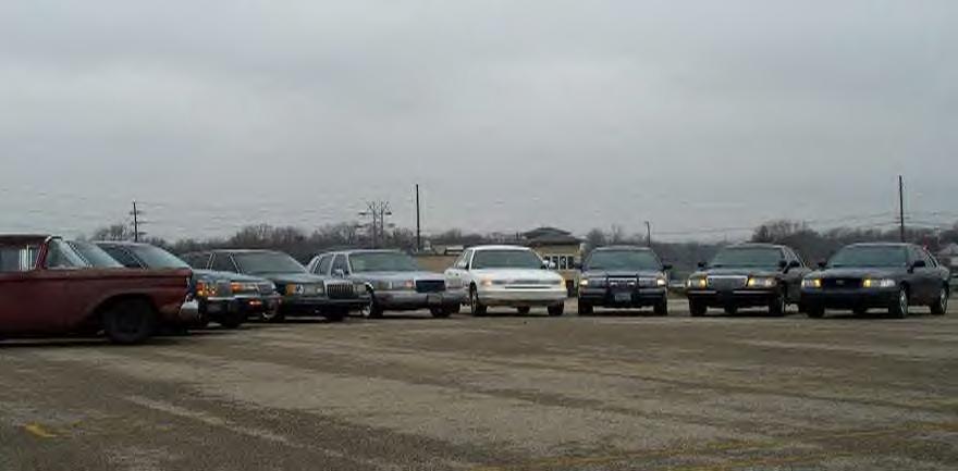 Curved row of parked sedans with headlights on, rusty red 1950s Ford closest at left