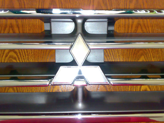 Chrome Mitsubishi emblem on a horizontal bar grille with wood grain background. Text 'SUPERMOTORS' visible