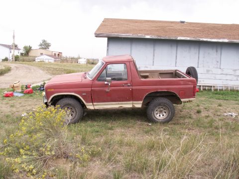 1986 Ford Bronco - Side view of a red Ford Bronco with a spare tire on the back, parked outdoors.