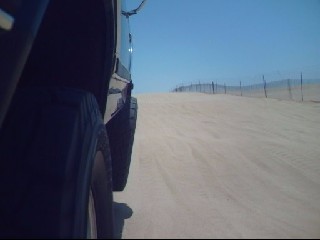 1984 Ford Bronco - The low-angle, side-view photo shows the rear tire and fender of a dark-colored SUV driving on a sandy track next to a fence under a clear…