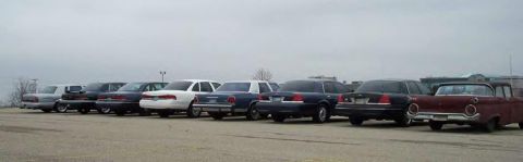 1988 Ford Crown Victoria - A collection of sedans and a vintage car are parked outdoors under an overcast sky.