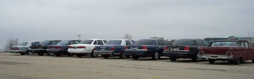 Row of full-size sedans and an older car parked in a gravel lot under gray sky