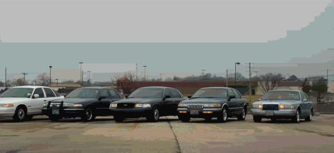 1988 Ford Crown Victoria - A lineup of Ford Crown Victorias, including a white model and four dark sedans, are parked outdoors under an overcast sky.