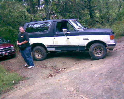 1988 Ford Bronco - A man stands next to a two-tone black and white Ford Bronco parked on a dirt road. A red car is parked to the left of the Bronco.
