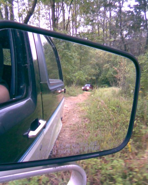 1988 Ford Bronco - A side view mirror reflects a red SUV driving on a dirt road through a wooded area. The mirror also shows text that reads "Objects in mirro…