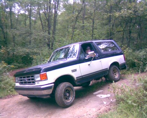 1988 Ford Bronco - A two-tone white and black Ford Bronco is driving on a dirt path through a wooded area. The vehicle is viewed from a front 3/4 angle.