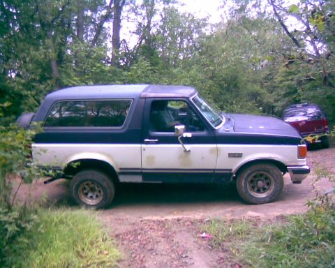 1988 Ford Bronco - A two-tone blue and white Ford Bronco is parked on a dirt road surrounded by trees. A red SUV is visible in the background.