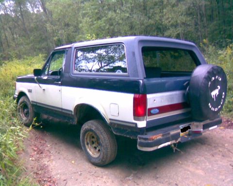 1988 Ford Bronco - The rear driver's side quarter view of a two-tone white and blue Ford Bronco is parked on a dirt road surrounded by trees. A spare tire cov…