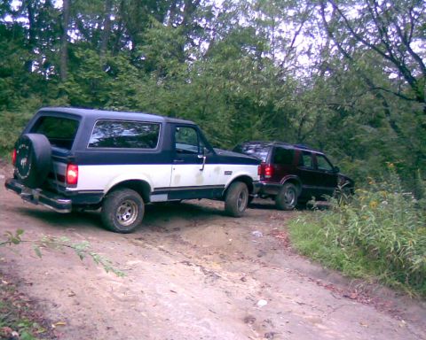 1988 Ford Bronco - A two-tone black and white Ford Bronco is parked on a dirt road next to a dark red SUV. The Bronco has a spare tire mounted on the rear.