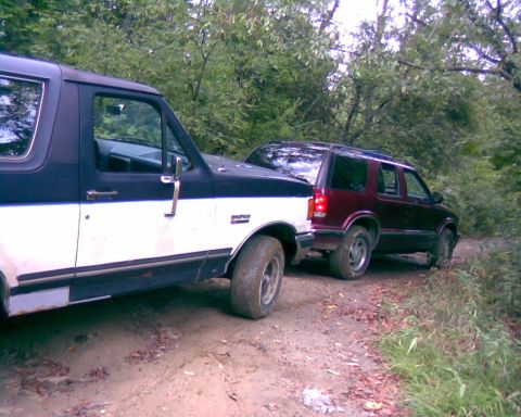1988 Ford Bronco - A two-tone black and white Ford Bronco is parked next to a dark red SUV on a dirt road surrounded by trees. The Bronco's front fender and d…