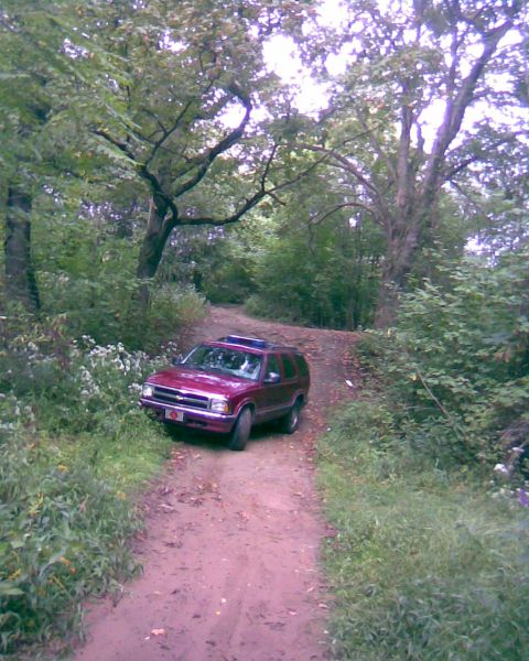 1988 Ford Bronco - A maroon SUV is parked on a dirt road surrounded by trees and foliage. The vehicle is facing towards the viewer with its front end slightly…
