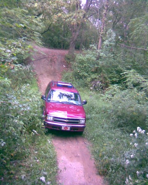 1988 Ford Bronco - A red SUV drives down a dirt path surrounded by lush green foliage. The vehicle has a sunroof and a chrome grille.