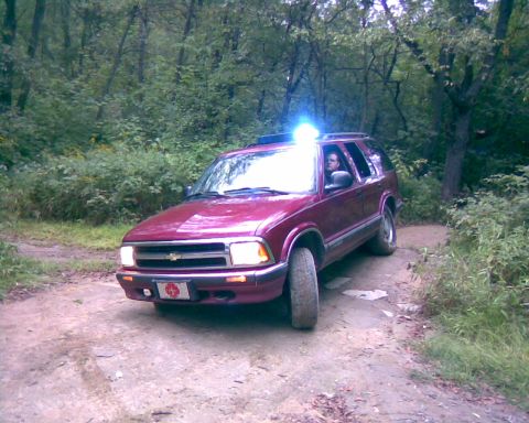 1988 Ford Bronco - A maroon SUV is parked on a dirt road in a wooded area. The vehicle has its headlights on and a blue light is visible on the roof.