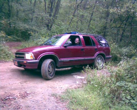 1988 Ford Bronco - A maroon SUV is parked on a dirt road in a wooded area. A person is visible in the driver's seat, looking towards the camera.