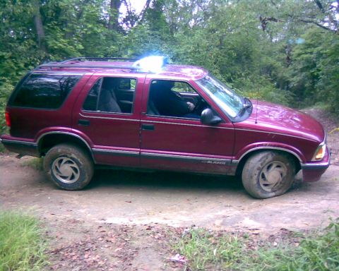 1988 Ford Bronco - A maroon SUV is parked on a dirt path in a wooded area. The front right tire appears to be flat.