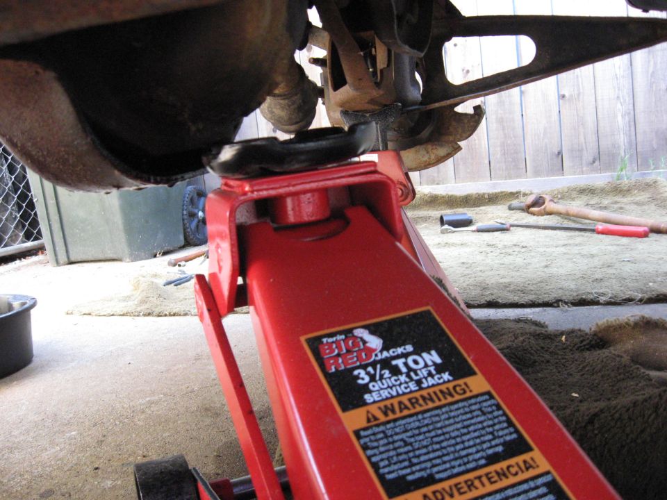 Red service jack lifting a vehicle, with tools and drive shaft on the ground.