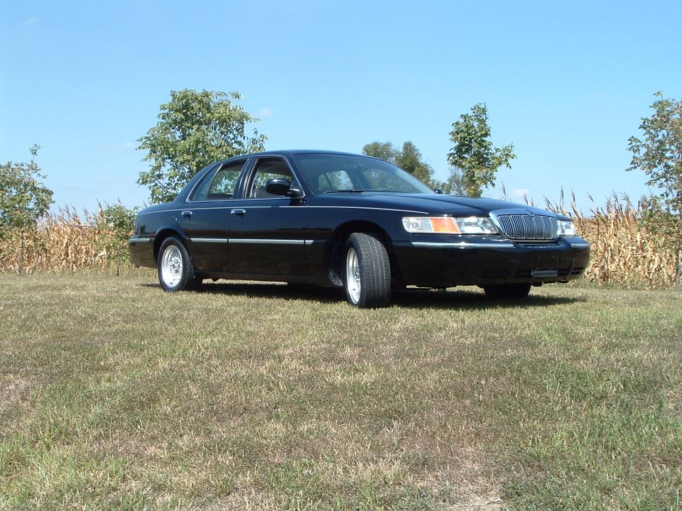 Black 2001 Mercury Grand Marquis sedan parked on grass with cornfield and trees behind.