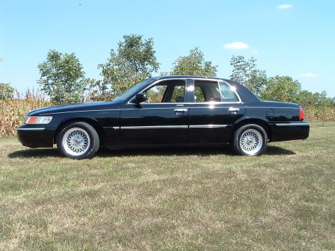 2001 Mercury Grand Marquis - The side profile of a black 2001 Mercury Grand Marquis is shown parked on a grassy lawn with trees and a cornfield in the background under…
