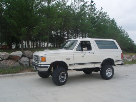 1987 Ford Bronco - A lifted, white 1987 Ford Bronco is parked on a concrete surface with a line of pine trees and large rocks in the background.