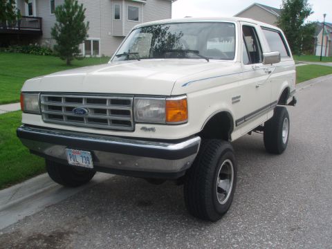 1987 Ford Bronco - A white 1987 Ford Bronco is parked on an asphalt street in front of a residential house. The vehicle has a chrome grille, aftermarket wheel…
