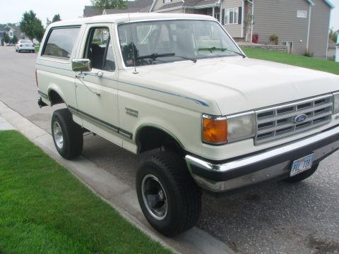 1987 Ford Bronco - A lifted, cream-colored 1987 Ford Bronco is parked on a residential street next to a curb and grass. The front of the vehicle is angled tow…