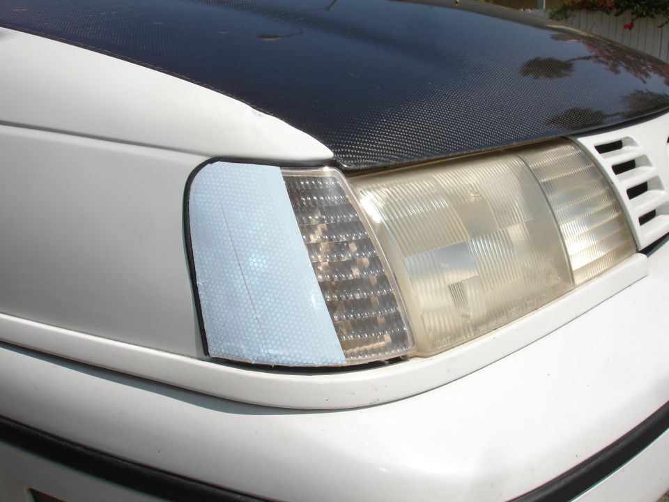 White car front corner with carbon fiber hood wrap, clear headlight, and blue reflective turn signal sticker.