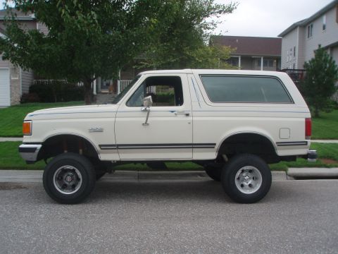 1987 Ford Bronco - The side view of a lifted, cream-colored 1987 Ford Bronco is parked on a residential street. The vehicle has blue pinstripes along its body…