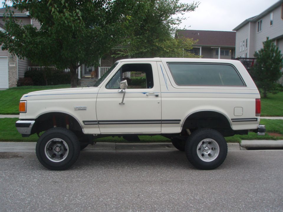 Lifted cream 1987 Ford Bronco with blue pinstripes and aftermarket wheels, parked on a street.