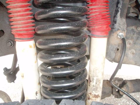 1987 Ford Bronco - A close-up view of the front suspension of a vehicle, featuring black coil springs and red-booted shock absorbers. A black hydraulic line i…