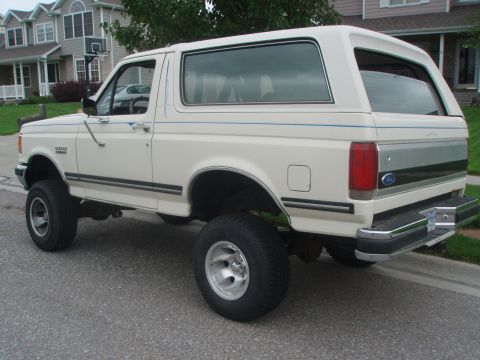 1987 Ford Bronco - A white 1987 Ford Bronco is parked on a residential street, viewed from the rear passenger side quarter angle. The vehicle has a lifted sus…
