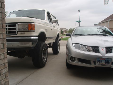 1987 Ford Bronco - A lifted, cream-colored 1987 Ford Bronco is parked next to a silver Pontiac Sunfire. The Bronco is viewed from the front-left, showing its…