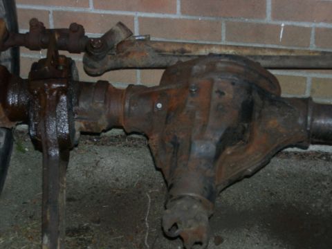 1996 Ford Bronco - A rusty, heavily used front axle assembly with steering components is shown resting on the ground against a brick wall.