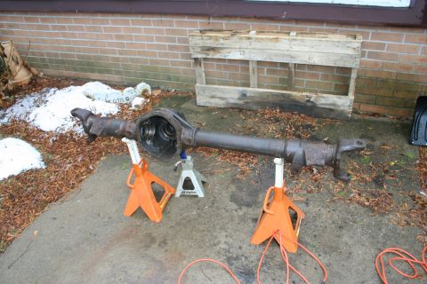 1996 Ford Bronco - A rear axle assembly is supported by three jack stands on a concrete driveway. The axle is rusty and appears to be from an older vehicle.