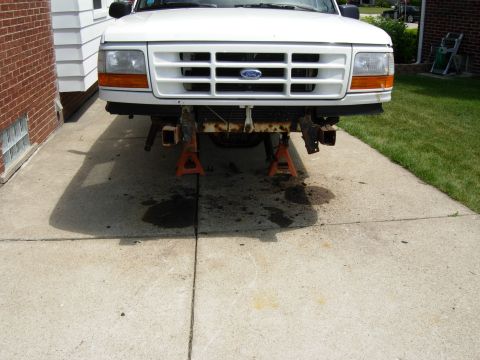 1996 Ford Bronco - The front end of a white Ford Bronco is raised on orange jack stands in a driveway. The grille, headlights, and bumper are visible, along w…
