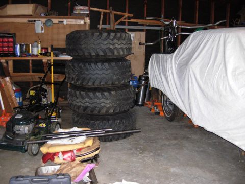 1996 Ford Bronco - A white car cover is draped over a vehicle, partially obscuring it. To the left, a stack of four large tires and a lawnmower are visible in…