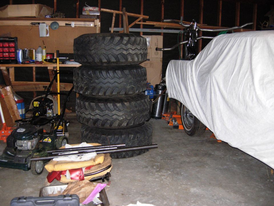 Vehicle covered in white sheet in a garage with tires and lawnmower nearby.