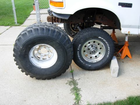 1996 Ford Bronco - Two large off-road tires are shown on a concrete surface in front of a white Ford Bronco that is supported by orange jack stands. The tire…