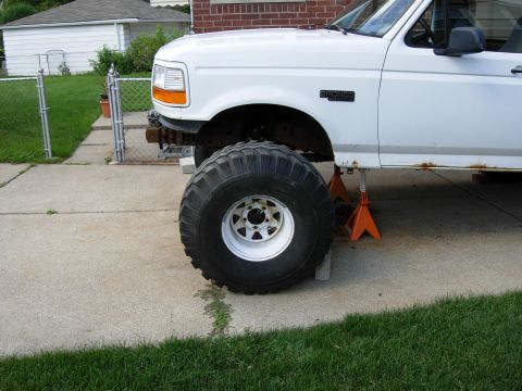 1996 Ford Bronco - A white Ford Bronco is jacked up on orange jack stands, revealing a large off-road tire and suspension components. The vehicle shows signs…