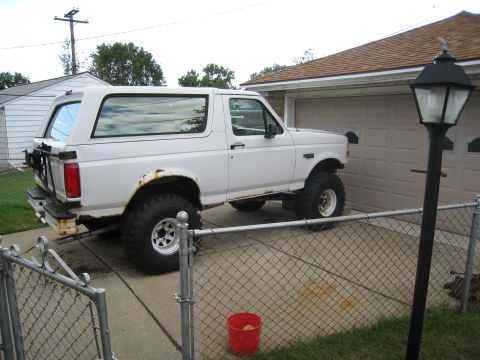 1996 Ford Bronco - A white 1996 Ford Bronco is parked in a driveway next to a garage. The vehicle has large off-road tires and visible rust on the rear fender.