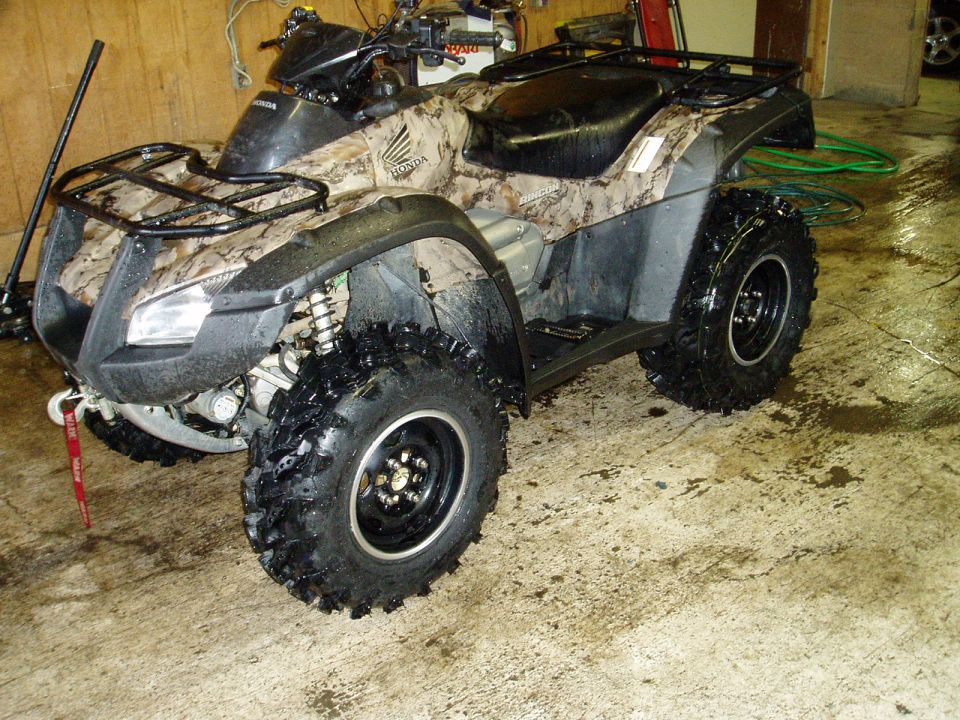 Camouflage Honda ATV parked on a wet concrete floor with a green hose.
