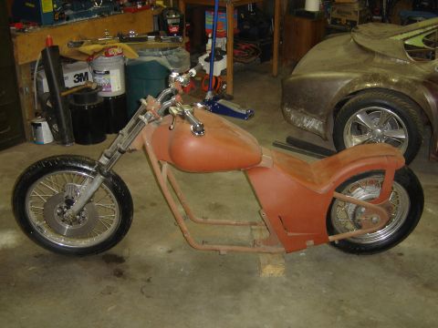 1994 Ford Probe - Unfinished custom motorcycle frame in a workshop setting, with a car body in the background.