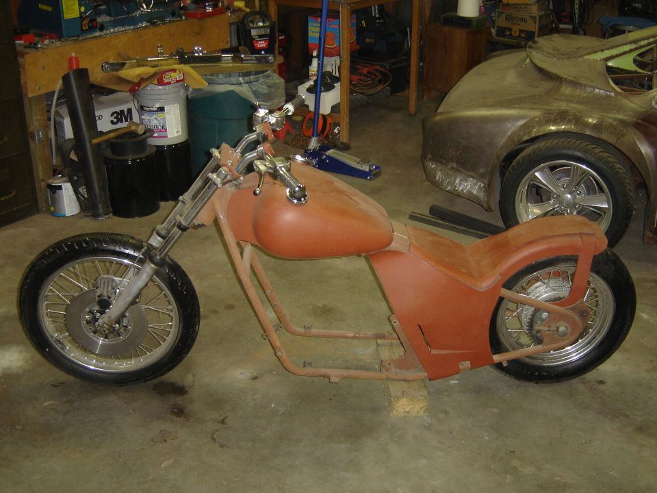 Custom motorcycle frame with fuel tank and seat pan, unfinished. Another vehicle body is visible in the background