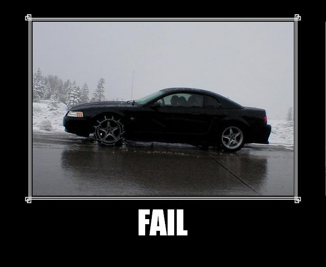Black coupe with snow chains on the front driver's side wheel, parked on a wet road in snowy conditions