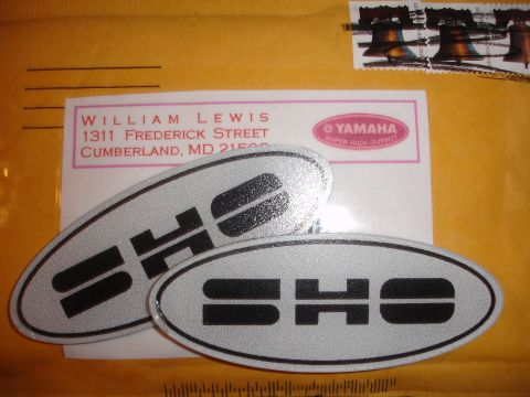 1990 Ford Taurus - Two silver oval SHO emblems with black lettering are shown on a yellow envelope with a return address.