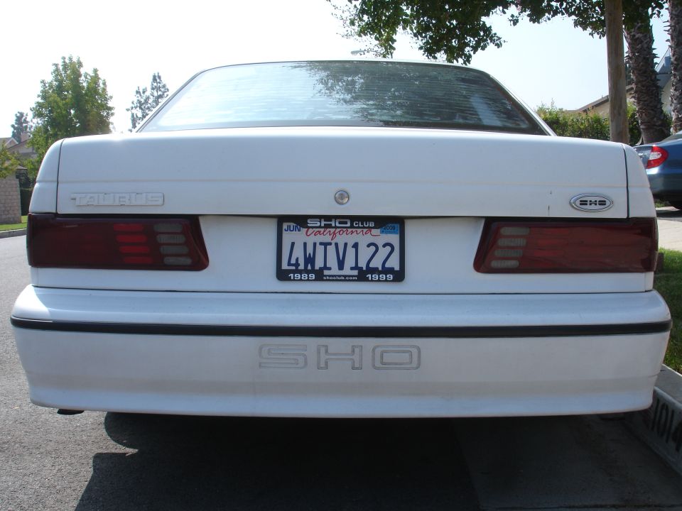 White Ford Taurus SHO with a California license plate that says 4WIV122. The plate also has '1989' and '1999' printed on it