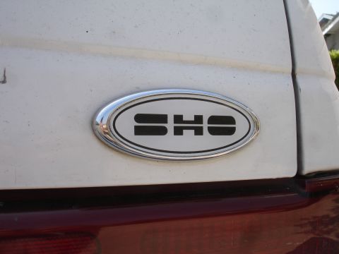 1990 Ford Taurus - Close-up of a white vehicle's rear section, showing an oval emblem with the letters 'SHO'.